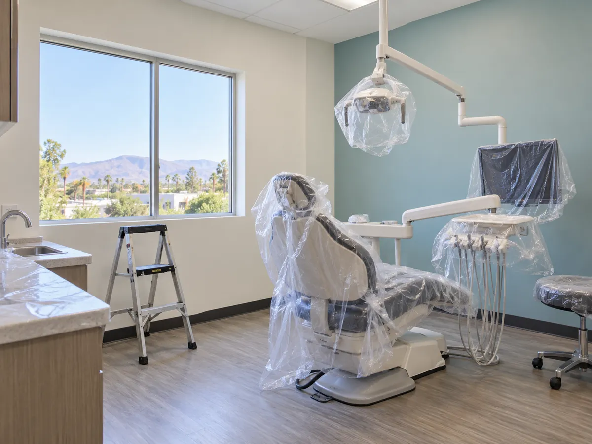A dental operatory with white walls and a blue-green accent wall, the chair and light covered in plastic sheeting, a step ladder by a window overlooking palms and mountains